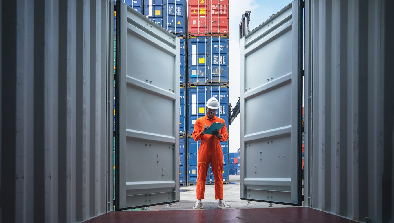 Man in work clothes taking notes while looking into empty container in container shipyard.