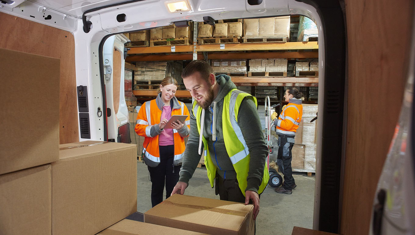 Warehouse worker loading delivery van while another takes inventory on tablet.