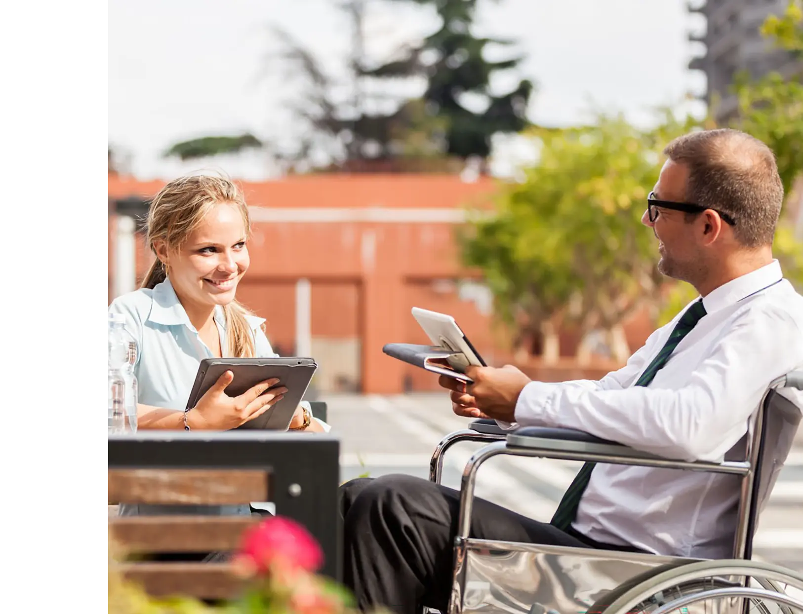 Woman on tablet talking to man with tablet in wheelchair.